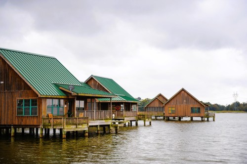 Wooden houses with green roofs on stilts over a calm lake, under a cloudy sky.