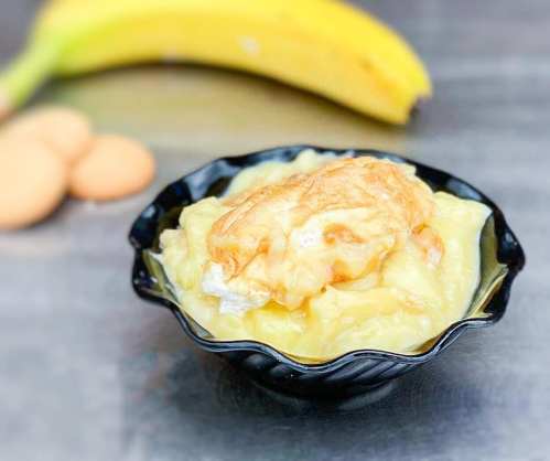 A black bowl filled with creamy banana pudding, with a banana and cookies in the background.