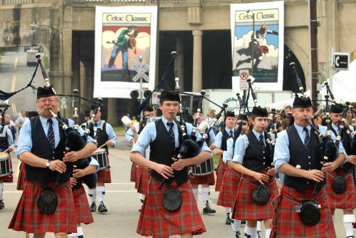 A group of musicians in kilts playing bagpipes and drums at a parade, with banners in the background.