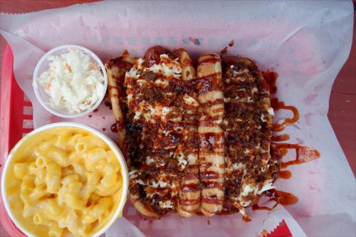 A red tray with three BBQ hot dogs topped with coleslaw and sauce, alongside a cup of macaroni and cheese.