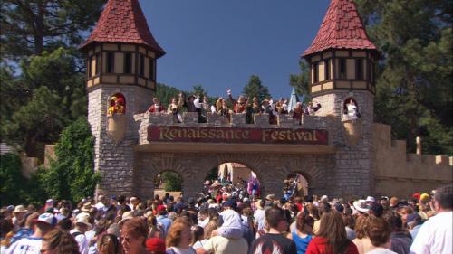 A large crowd gathers at a Renaissance Festival entrance, featuring towers and performers above a sign.