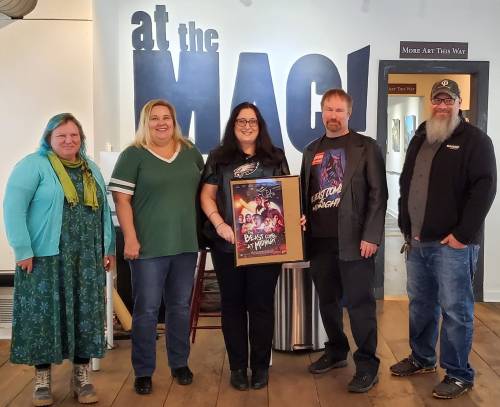 Five people stand together in an art gallery, holding a framed poster, with a sign reading "at the MAC" in the background.