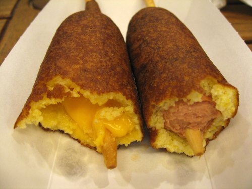 Two corn dogs on a white paper tray, one filled with cheese and the other with a hot dog, both partially bitten.