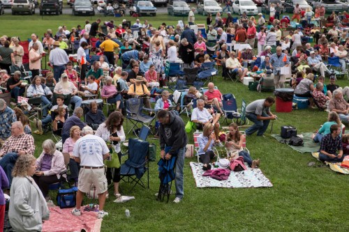 A large crowd of people sitting on chairs and blankets in a grassy area, enjoying an outdoor event.