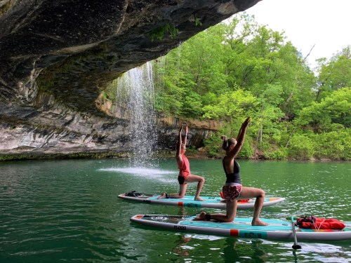 Two people practicing yoga on paddleboards under a rocky overhang with a waterfall in a lush green setting.