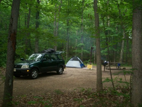 A green SUV parked near a tent in a wooded campsite, with a picnic table and campfire setup nearby.