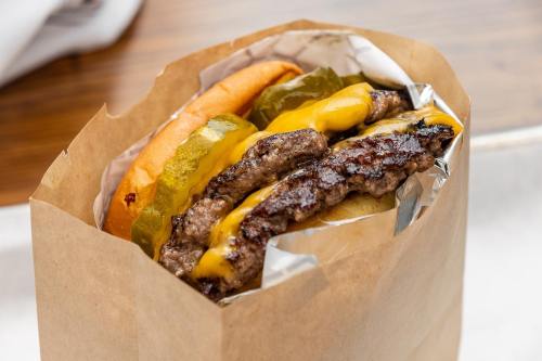 A close-up of a burger in a paper bag, featuring two beef patties, melted cheese, pickles, and a soft bun.