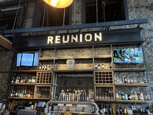 A bar with a "REUNION" sign, shelves of liquor, and a TV displaying a sports game in a rustic setting.