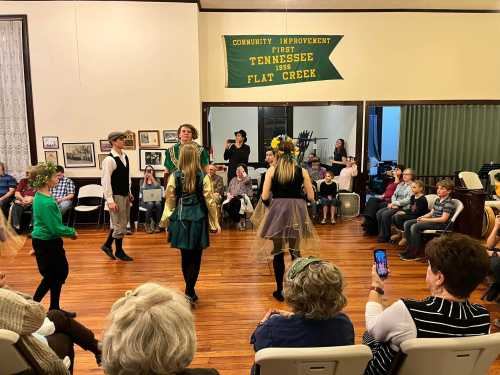 A group of dancers perform in a community hall, with an audience watching and a banner in the background.