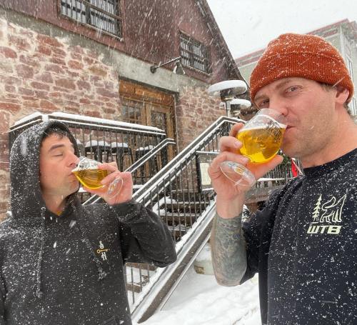 Two people in winter attire enjoy beers outside in a snowy setting, with a rustic building in the background.
