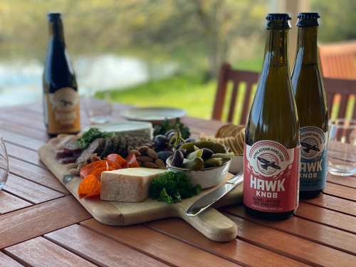A wooden table set with a charcuterie board, two bottles of Hawk Knob cider, and a scenic outdoor background.