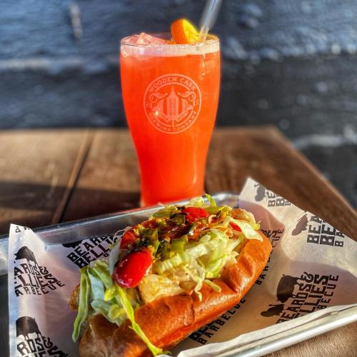 A colorful hot dog topped with lettuce, tomatoes, and pickles, alongside a vibrant drink in a branded glass.