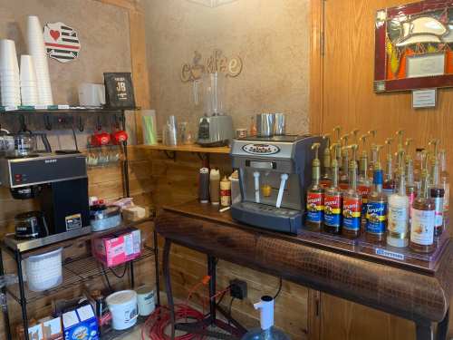 A cozy coffee shop corner with a coffee machine, syrup dispensers, cups, and various drink supplies on shelves.