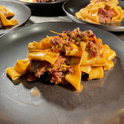 A close-up of a plate featuring pappardelle pasta topped with meat and a colorful garnish.
