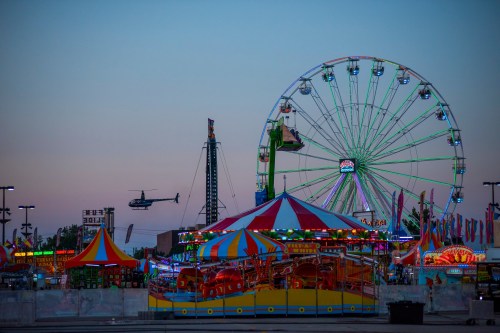 A vibrant carnival scene at dusk featuring a Ferris wheel, rides, and a helicopter flying overhead.