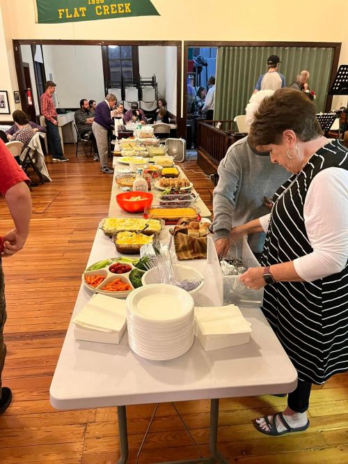 A buffet table filled with various dishes, with people serving food in a community hall setting.