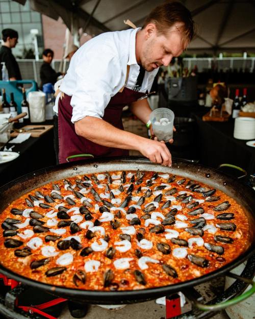 A chef prepares a large paella with seafood at an outdoor event, focusing on the dish in a large pan.