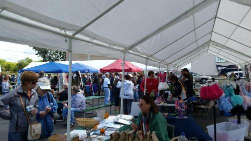 A bustling outdoor market with vendors under tents, showcasing clothing and crafts, and visitors browsing the stalls.
