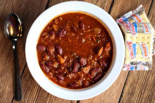 A bowl of chili with beans and meat, accompanied by a spoon and a pack of crackers on a wooden surface.
