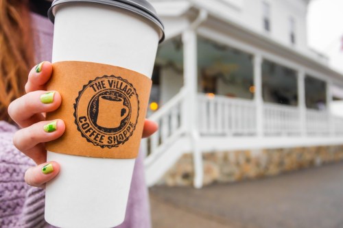 A person holds a coffee cup with a logo from "The Village Coffee Shoppe" in front of a white building.