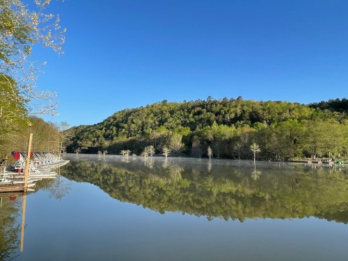 A serene lake reflecting lush green hills under a clear blue sky, with boats lined along the shore.