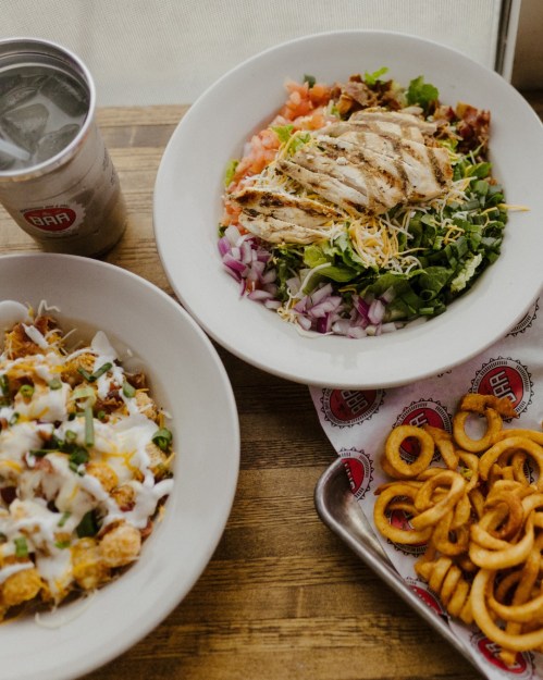 A bowl of salad with grilled chicken, tomatoes, and cheese, alongside a dish of loaded nachos and curly fries.