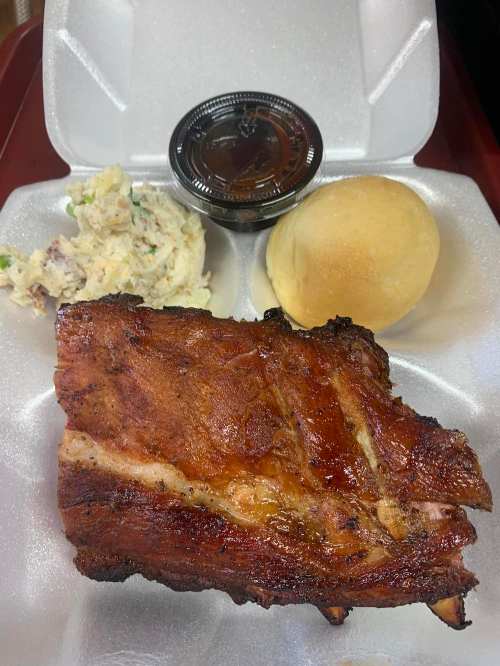A takeout container with a piece of grilled meat, a roll, coleslaw, and a small cup of sauce.