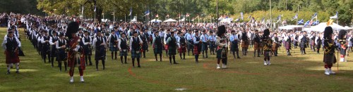A large gathering of pipers in traditional attire performing outdoors, surrounded by a crowd and flags.