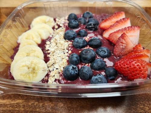 A clear container filled with acai bowl topped with banana slices, strawberries, blueberries, and granola.