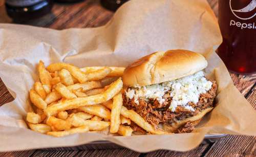 A pulled pork sandwich topped with coleslaw, served with a side of crispy French fries in a paper-lined basket.