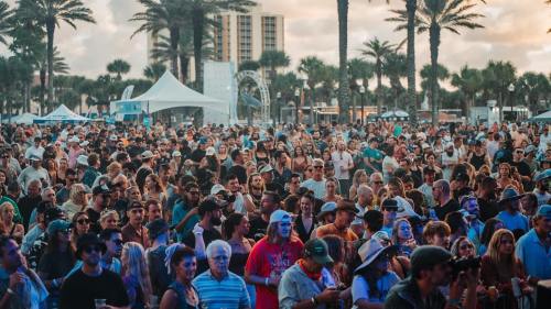 A large crowd of people at an outdoor event, with palm trees and tents in the background under a cloudy sky.
