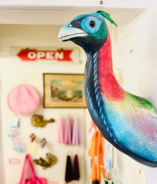 A colorful bird sculpture hangs on a wall, with an "OPEN" sign and various items in the background.