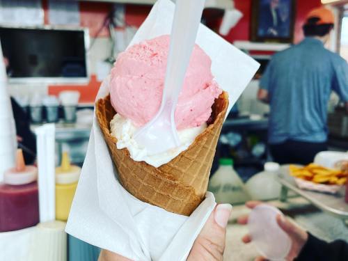 A hand holds a waffle cone filled with pink and white ice cream, with a busy ice cream shop in the background.