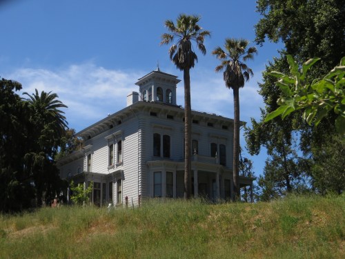 A vintage two-story house with a tower, surrounded by palm trees and lush greenery under a blue sky.