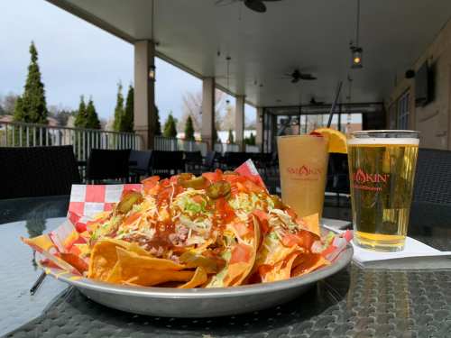 A plate of nachos topped with lettuce, tomatoes, jalapeños, and a side of beer and a cocktail on a patio table.