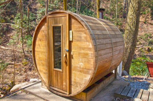 A wooden barrel-shaped sauna nestled in a forested area, featuring a door and chimney.