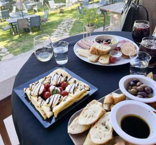 A table set with a variety of dishes, including a flatbread with tomatoes, olives, cheese, and drinks, in a garden setting.