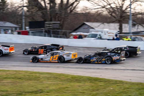 Five race cars compete on a track, with one car in the foreground making a sharp turn. Trees and spectators in the background.
