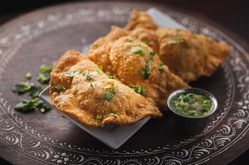 Three golden-brown samosas garnished with cilantro, served on a plate with a small bowl of green chutney.