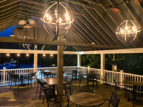 A spacious outdoor dining area with wooden tables, black chairs, and decorative lighting under a sloped roof at dusk.