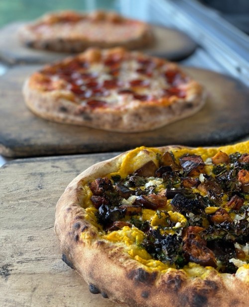 Three pizzas on wooden boards: one with a golden crust topped with vegetables, and two others in the background.