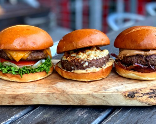 Three gourmet burgers on a wooden board: one with cheese and lettuce, one with a crispy topping, and one with peanut butter.