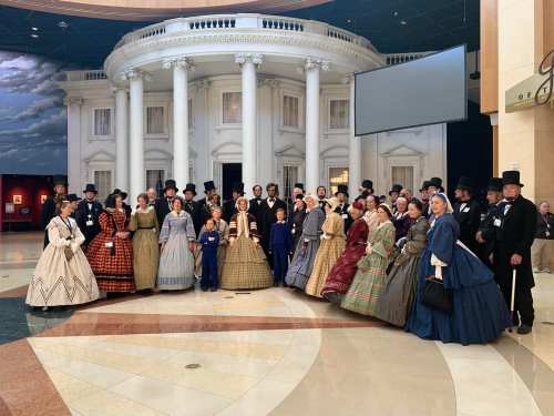 A large group in 19th-century attire poses in front of a replica of a historic building.