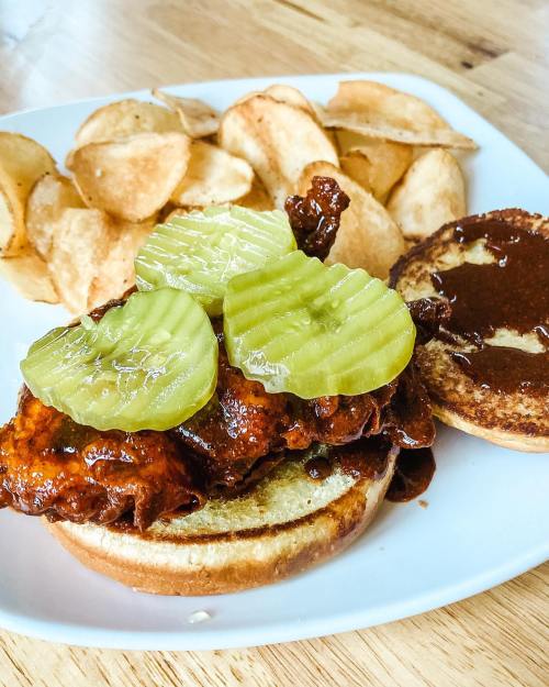 A close-up of a spicy chicken sandwich topped with pickles, served with a side of crispy potato chips on a white plate.