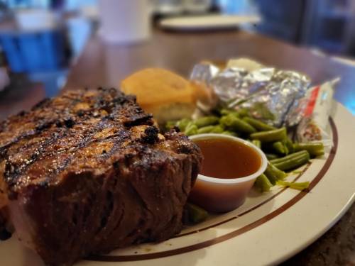 A plate featuring a grilled steak, green beans, cornbread, and a small cup of sauce.