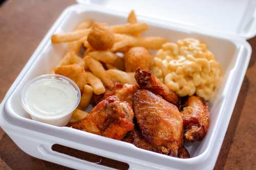 A takeout container with crispy chicken wings, fries, macaroni and cheese, and a side of ranch dressing.