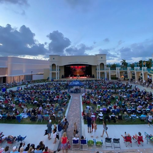 A large crowd gathers on a lawn for an outdoor concert at dusk, with a stage illuminated in the background.