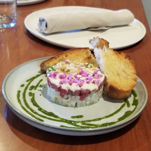 A plate with layered sushi topped with flowers, served with toasted bread and a green sauce drizzle.