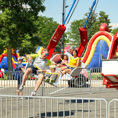 A man swings with a child at a fair, while a woman and another child enjoy a nearby ride. Colorful inflatables in the background.