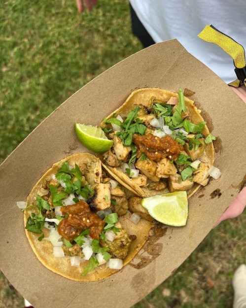 Two tacos filled with grilled chicken, onions, cilantro, and salsa, served with lime wedges on a brown paper tray.